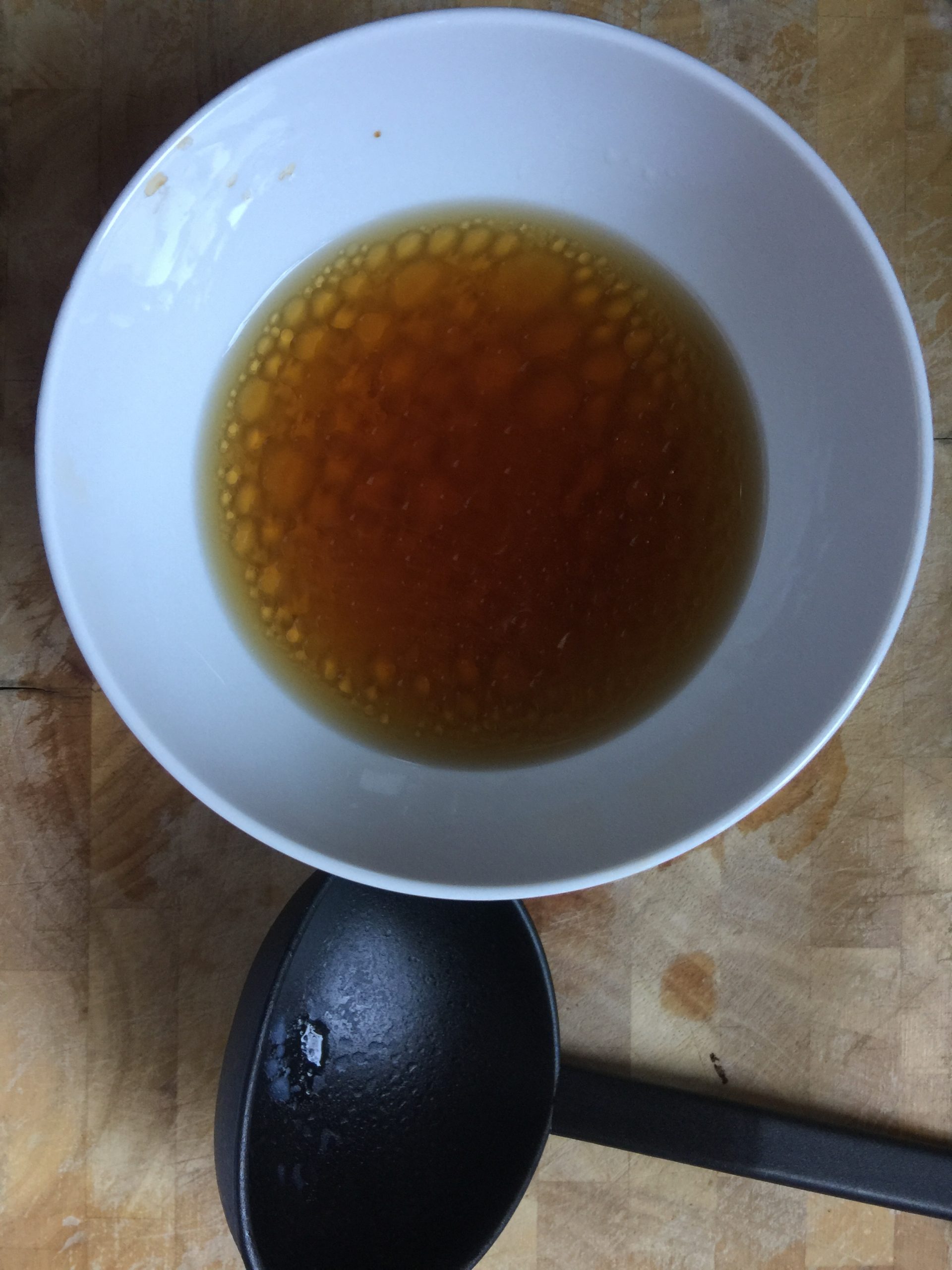 A white bowl of 1 tablespoon of soy sauce and sesame oil, diluted with one soup spoon of the boiling water, the soup spoon lying on the side.