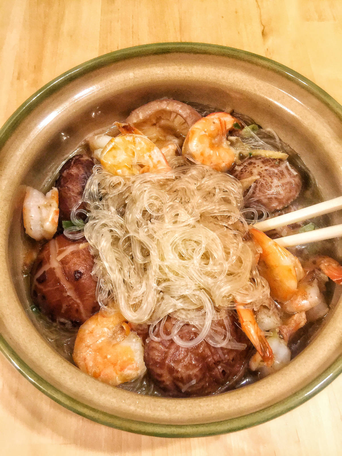 Cooked vermicelli noodles, shelled shrimps and shiitake mushrooms in a clay pot with a pair of chopsticks stick to the pot