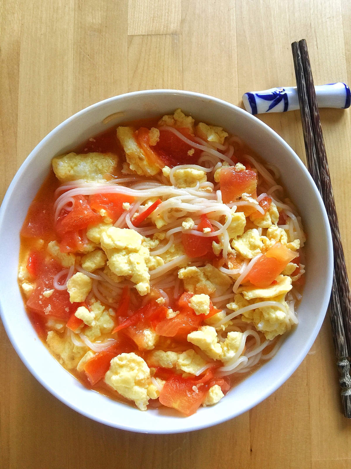 Overhead view of finished tomato egg noodle soup in a white bowl on a wooden table next to a pair of brown bamboo chopsticks resting on a ceremic chopstick holder