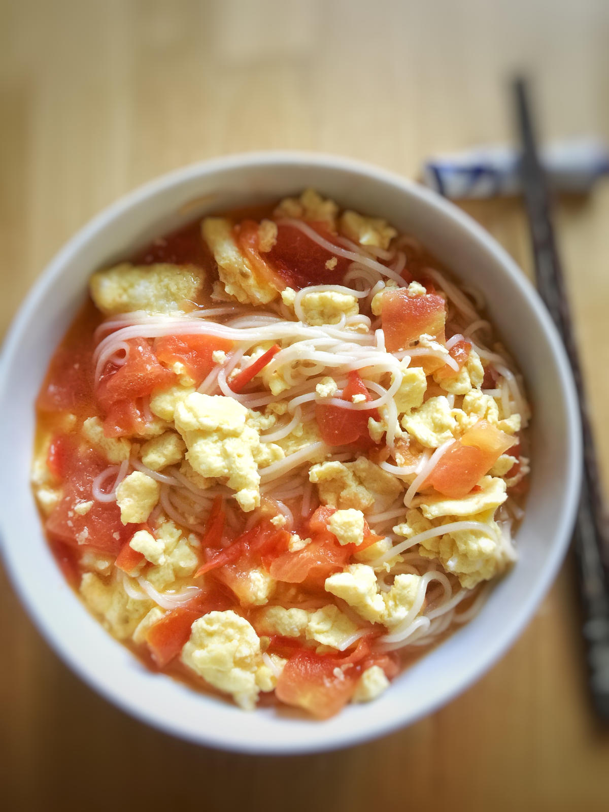 tomato egg noodle soup in a white bowl on a wooden table with a pair of chopsticks resting on a holder