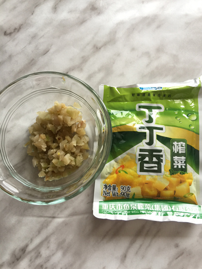 A small transparent bowl with chopped Chinese preserved vegetable (zha cai) next to a package of the preserved vegetable.
