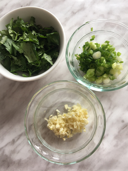 Overhead view of 3 small transparent bowls with chopped coriander, minced garlic and finely chopped scallion.