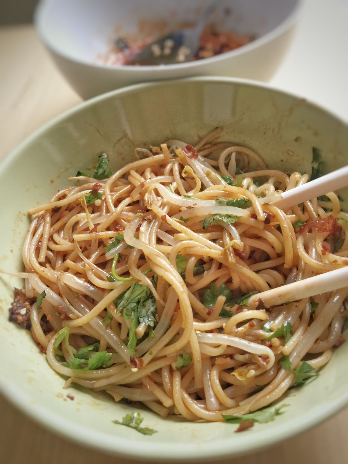Close up view of finished spicy cold noodles in a green bowl with a pair of ivory chopsticks. A white bowl of leftover sauce behind the green bowl