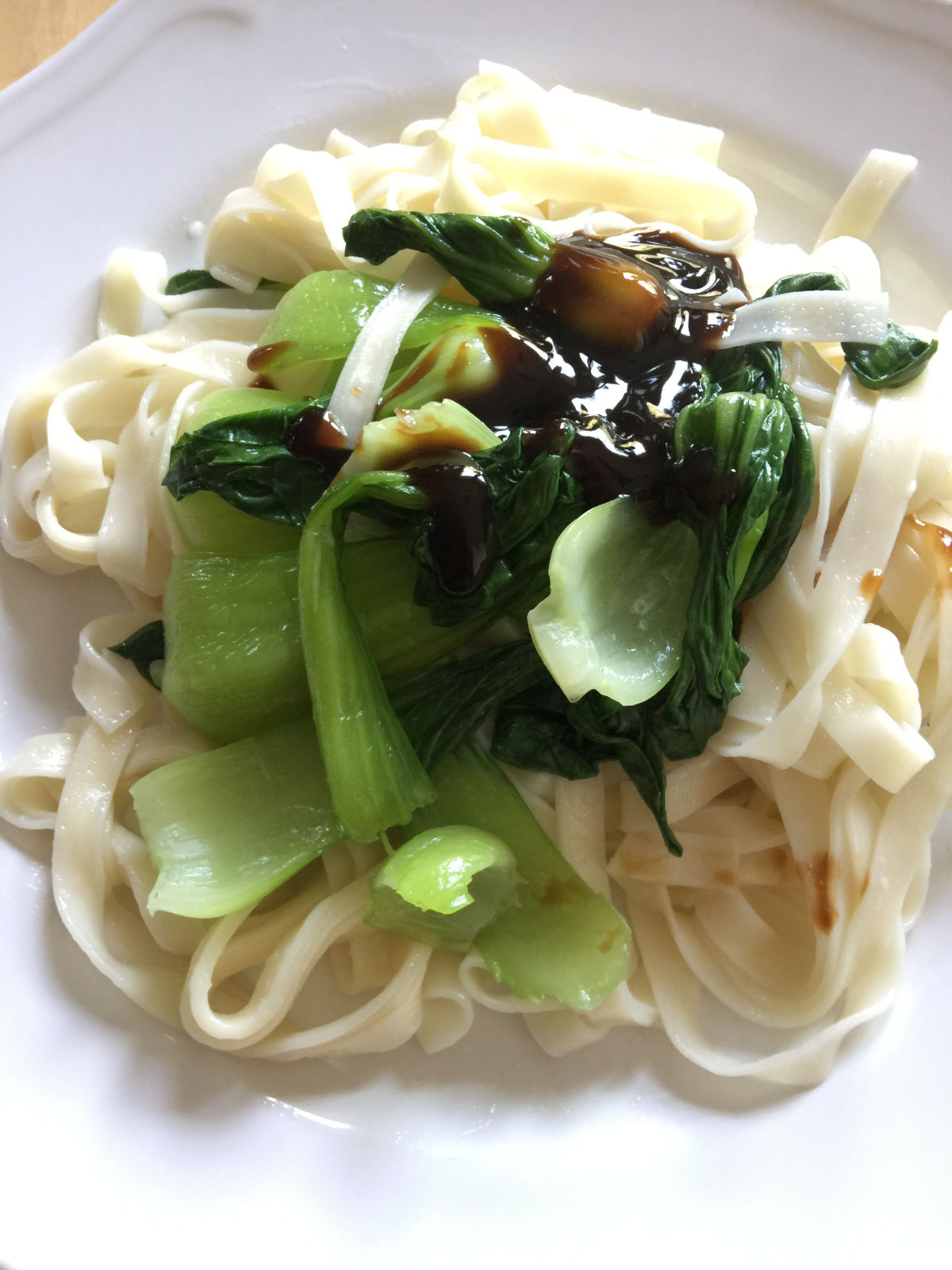 Close-up view of cooked noodles and bok choy placed on a white plate with oyster sauce on top.