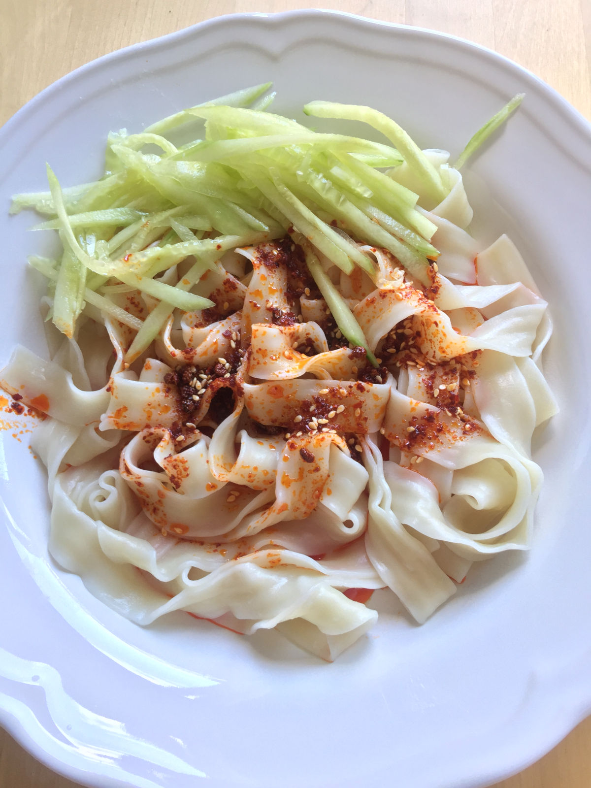 Overhead view of cooked noodles placed on a white plate with cucumber strips, chili oil and seasonings on top.