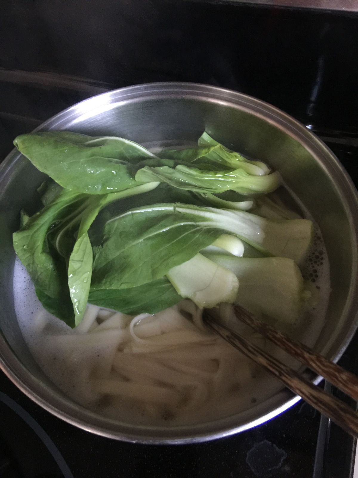 Overhead view of a large pot with boiling water, noodles and few plants of bok choy.