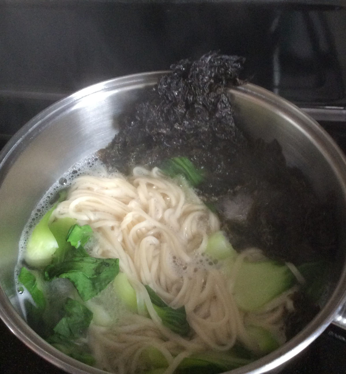 Noodles, bok choy and seaweed cooking in boiling water in the pot.
