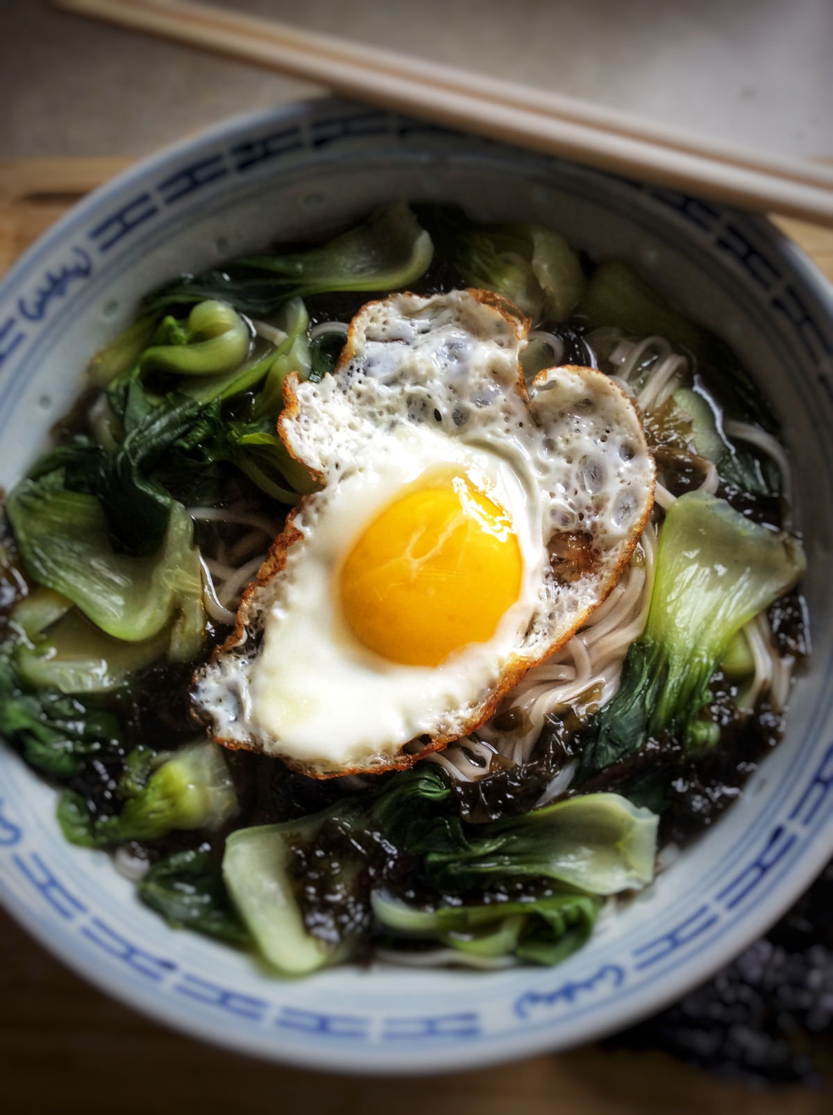 Close up view of cooked vegetable noodle soup in a big bowl with a fried egg on top and a pair of chopsticks resting on the bowl.