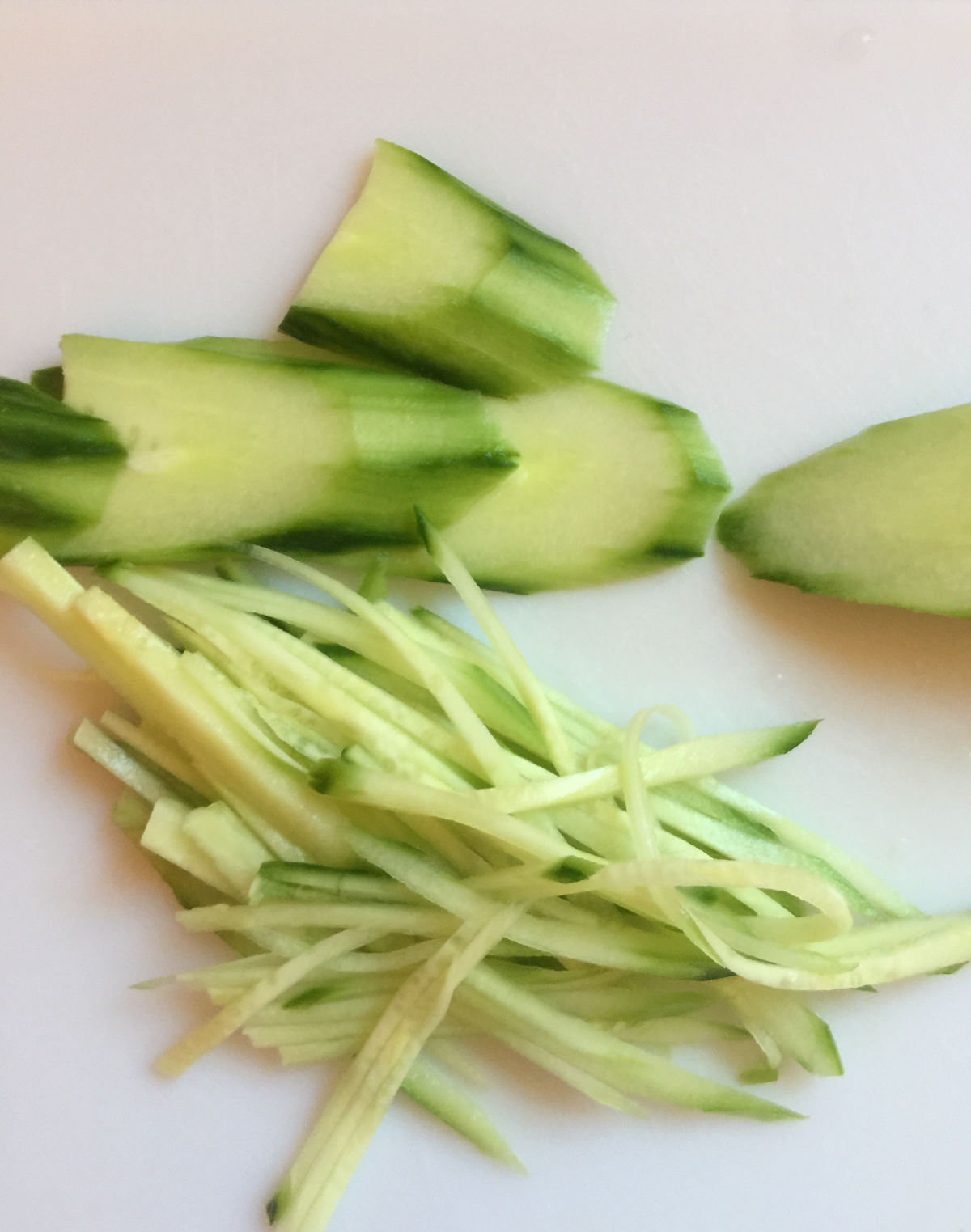 Peeled English cucumber cut into slices and thin matchstick-sized strips on a white cutting board.
