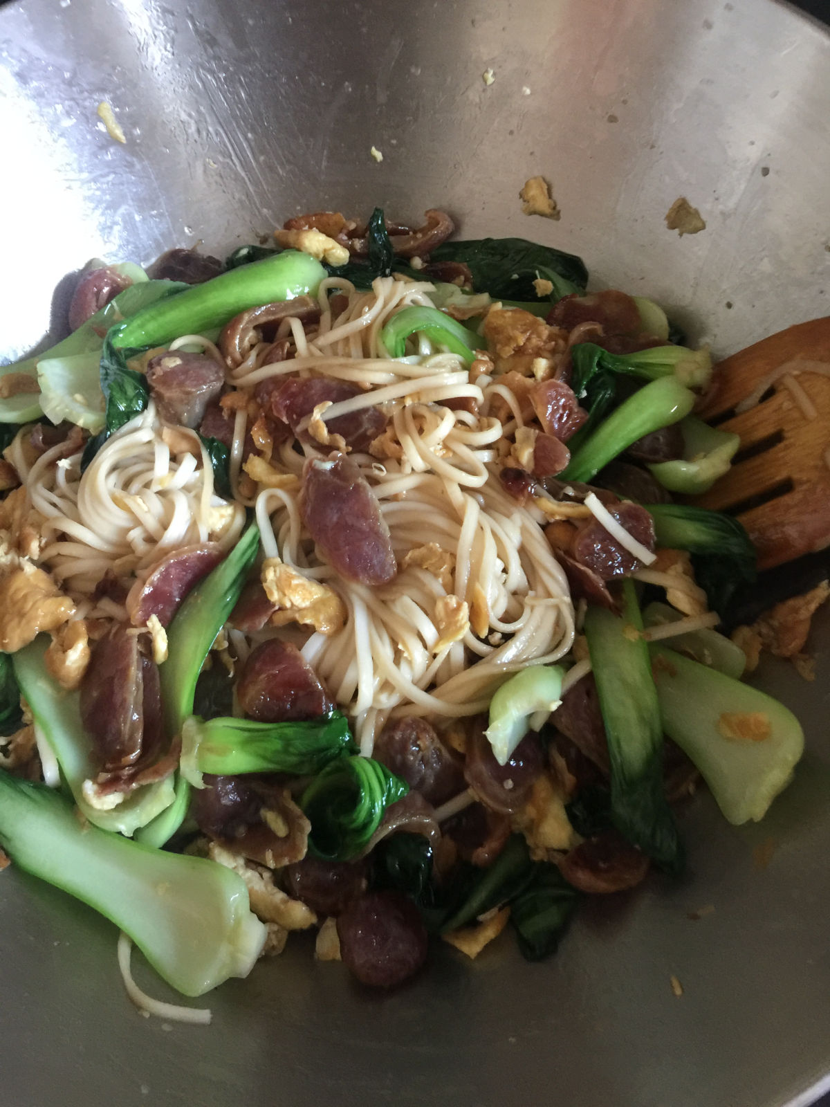 Noodles cooked with bok choy, eggs and sausages in wok with wooden spatula on the side.