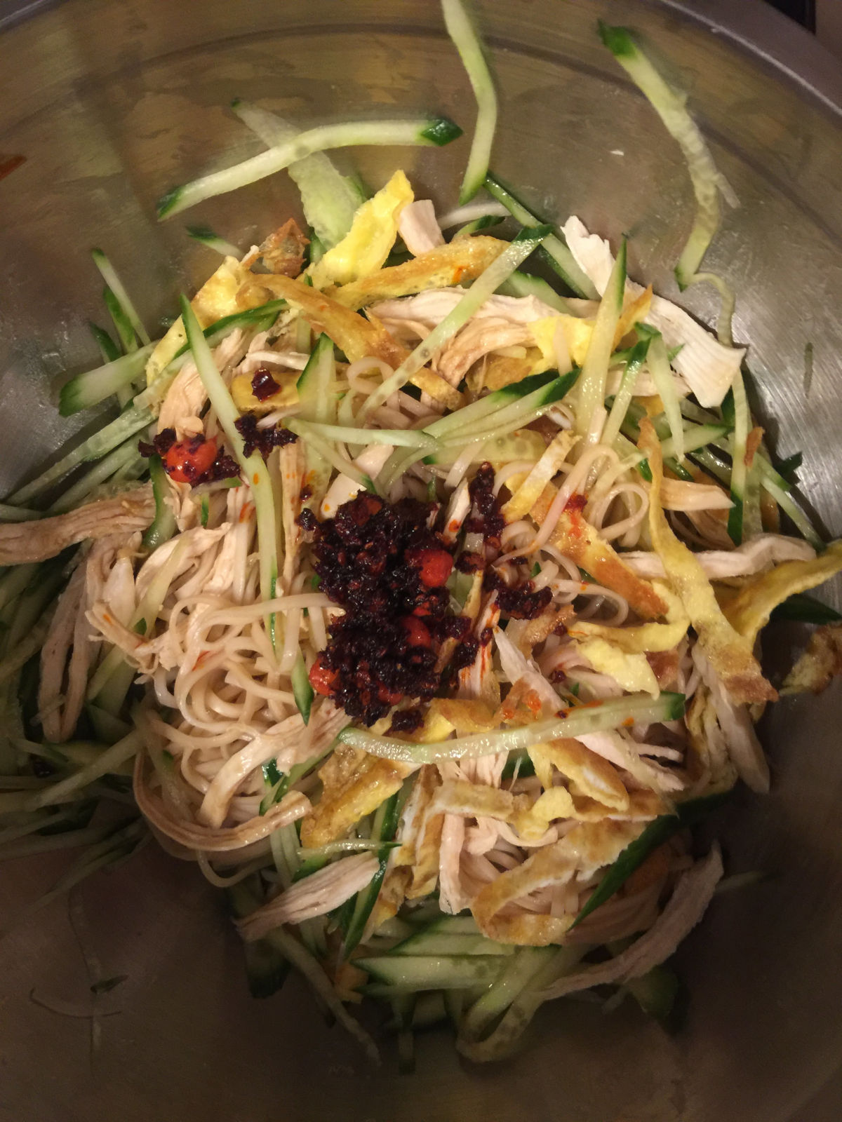 Close-up view of a large bowl with cucumber strips, shredded chicken breast, egg strips, cooked noodles and LaoGanMa chili sauce.