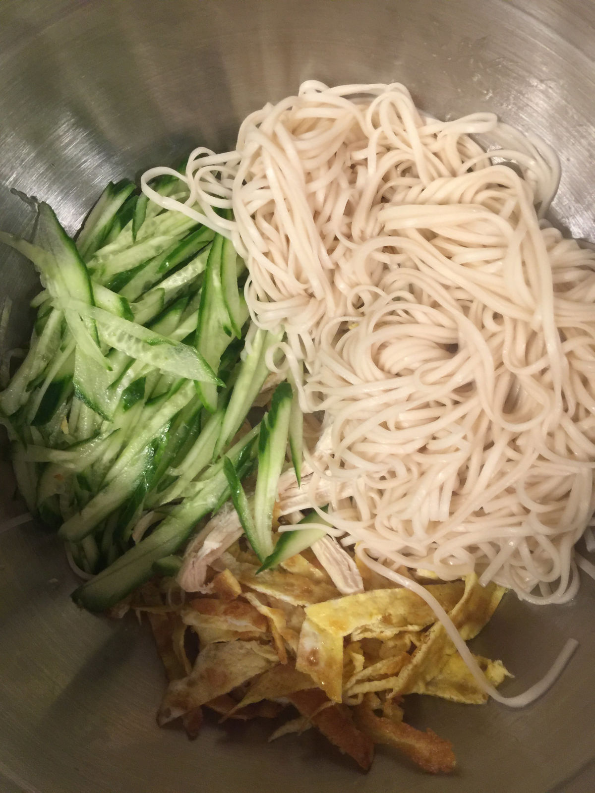 Overhead view of a large bowl with cucumber strips, shredded chicken breast, egg strips and cooked noodles.