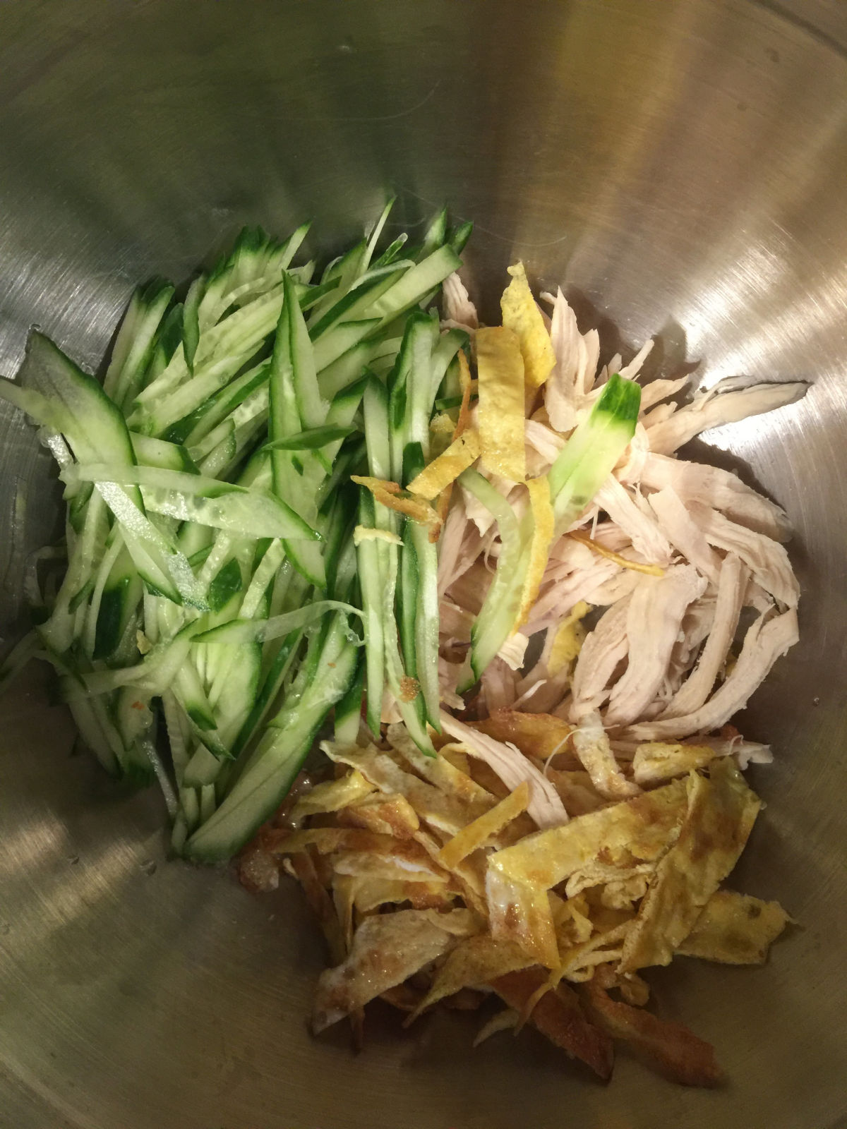 Overhead view of a large bowl with cucumber strips, shredded chicken breast, and egg strips.