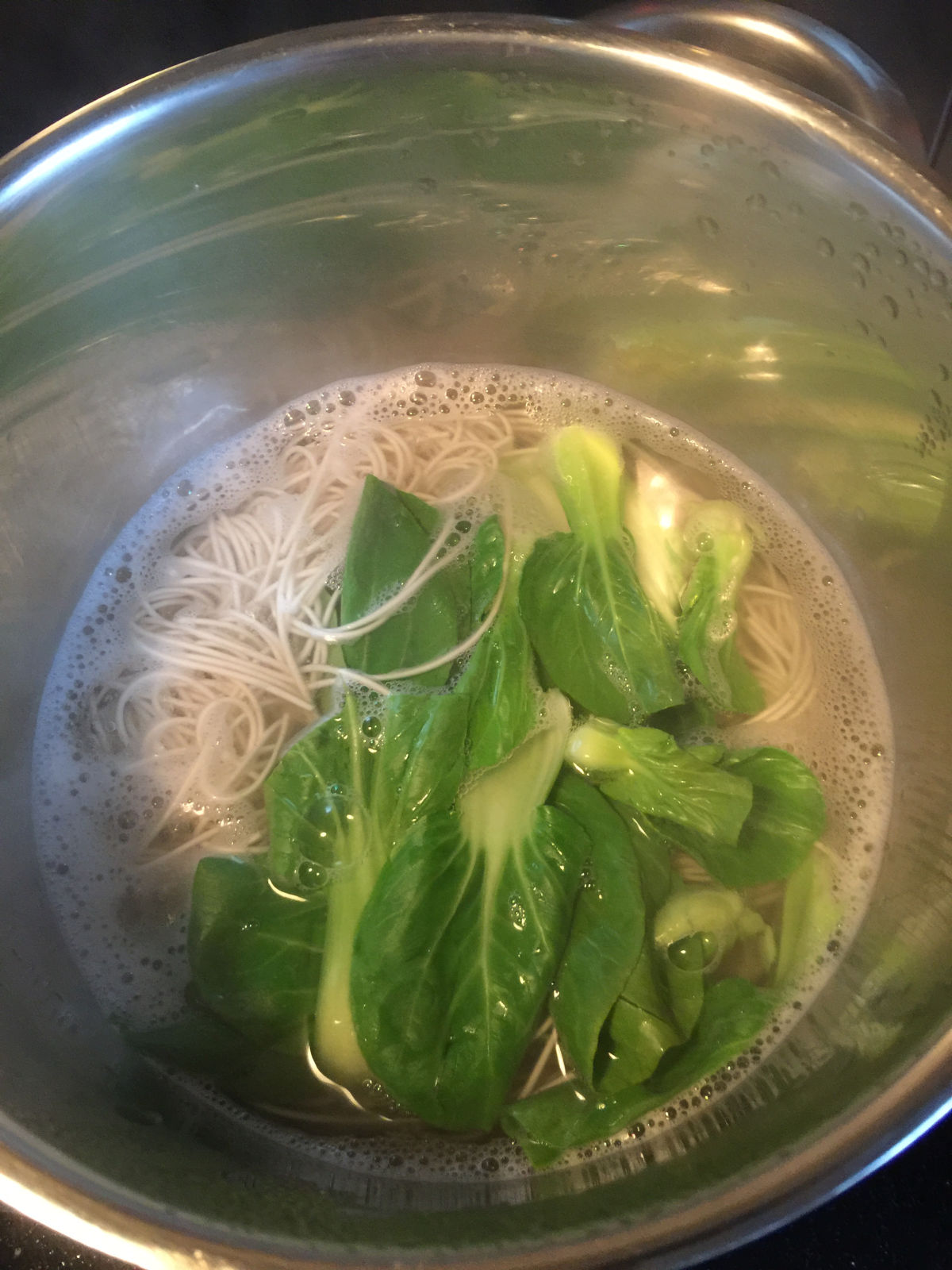 Overhead view of boiling noodles and bok choy in a stainless steel pot.
