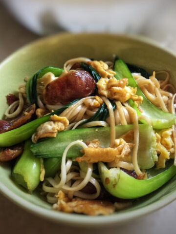 A close-up view of a bowl of cooked sausage and egg stir fried noodle.