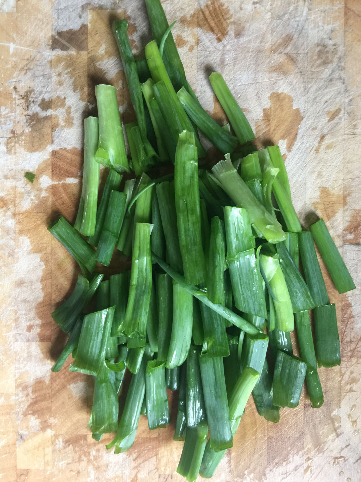 A bunch of 2-inch long pieces of spring onions (green part) on a wooden cutting board.