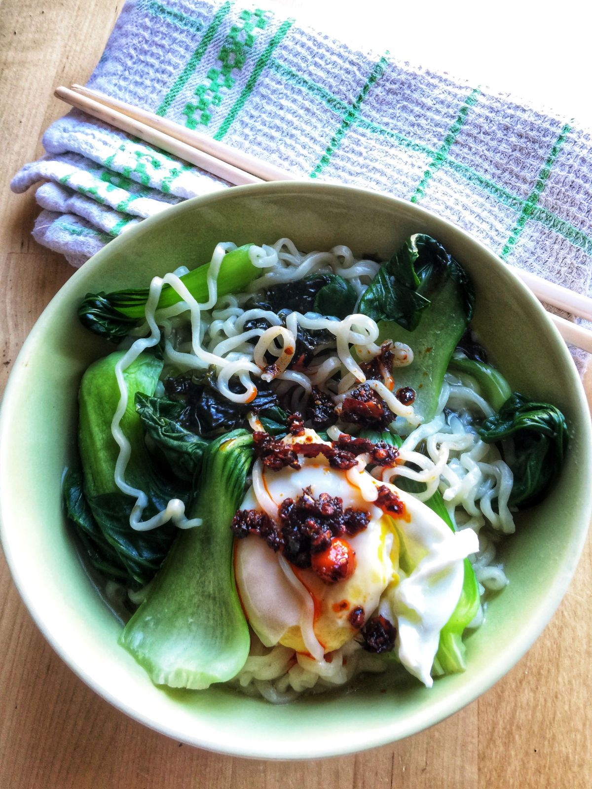 Overhead view of cooked instant noodle soup in a green bowl with 1 teaspoon LaoGanMa chili crisp on top, next to a pair of chopsticks on the table.