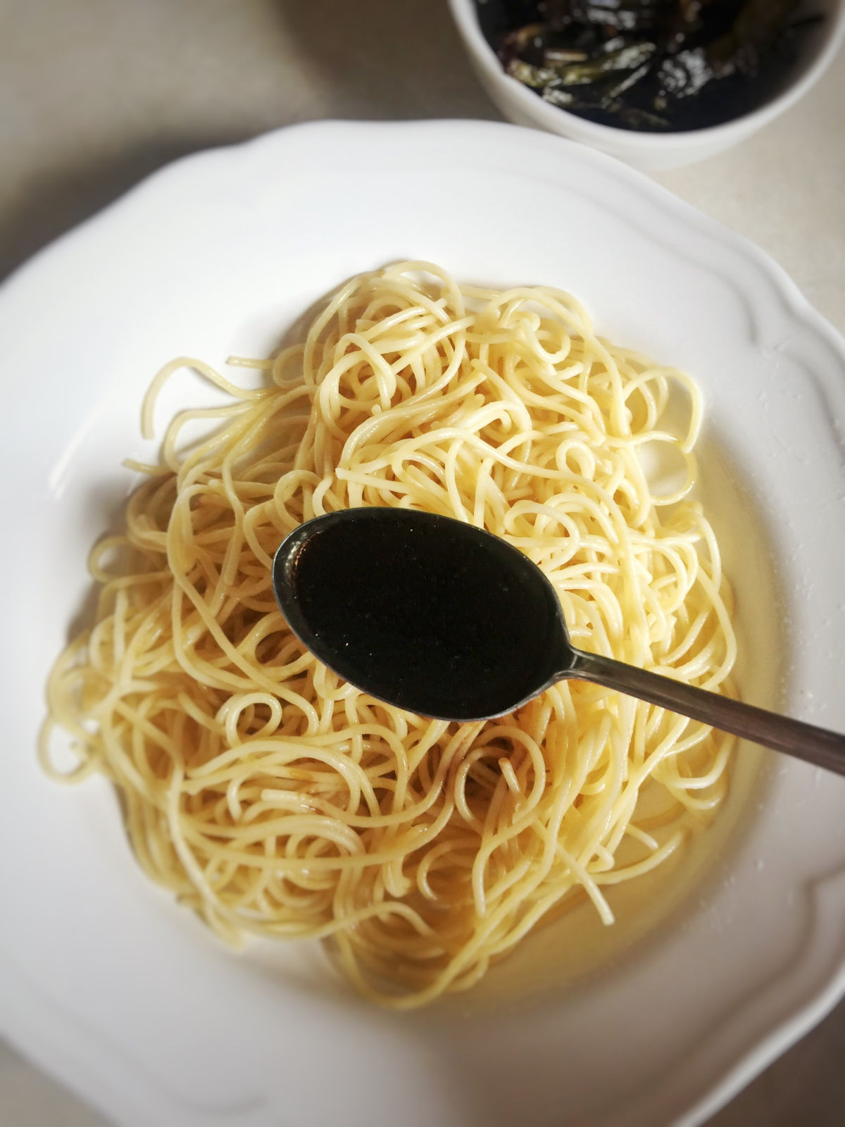 Overhead view of a white plate with drained pasta next to the cooked scallions placed in a small white bowl. A spoon of scallion oil to be added to the pasta.