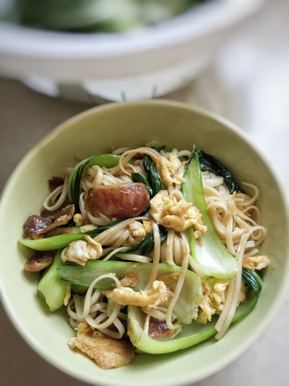 Close up view of cooked sausage and egg stir fried noodle, with a plastic basket of bok choy in the background.