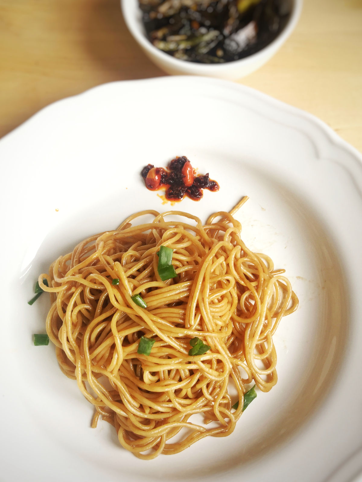 Overhead view of a white plate full of scallion oil pasta topped with finely chopped scallion and a teaspoon of Lao Gan Ma chili crisp on the side. Next to the cooked scallions placed in a small white bowl.