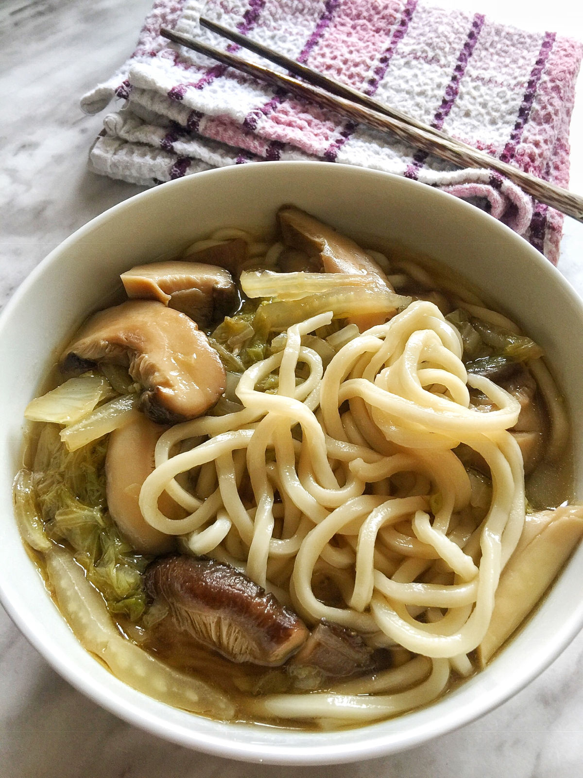 A close-up view of a bowl of cooked vegetable noodle soup with a pair of chopsticks on top of dishcloths next to the bowl.