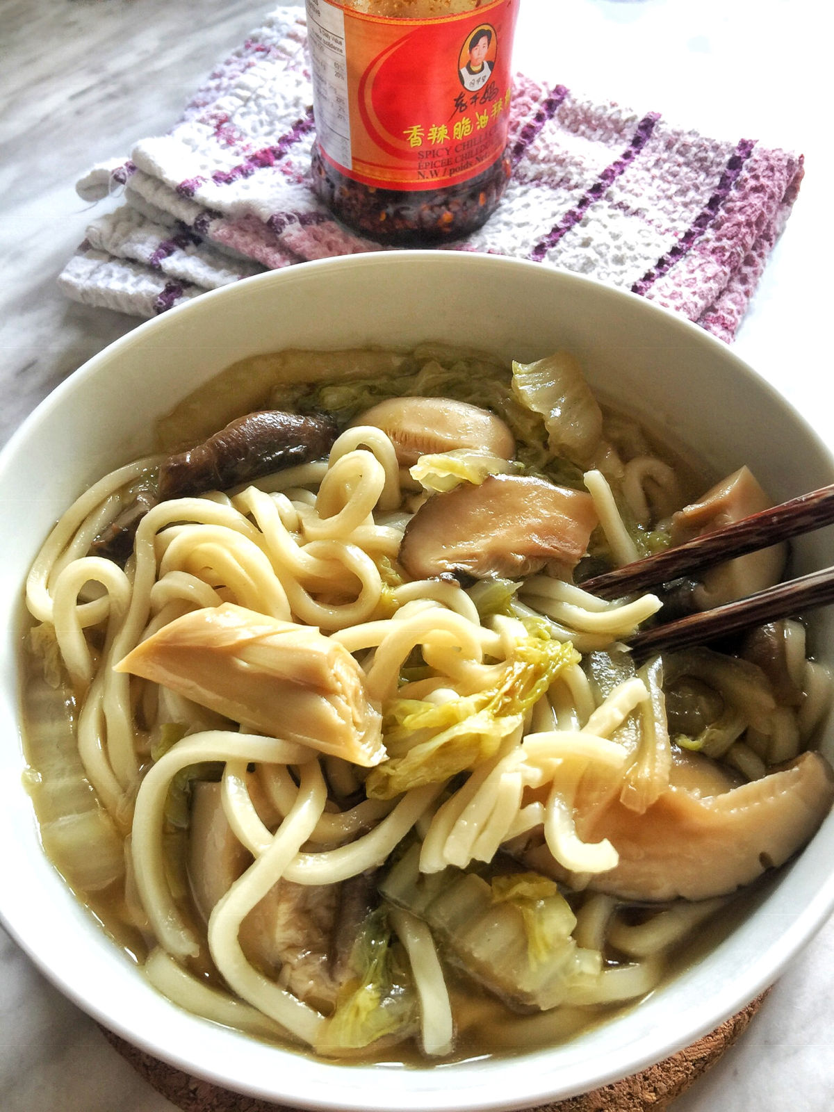 A close-up view of a bowl of cooked vegetable noodle soup with chopsticks on the side, a jar of Lao Gan Ma chili crispy next to the bowl.